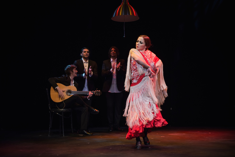 Pericet in a red dress with a white shawl dancing flamenco while two male singers and male guitarist accompany her in the background